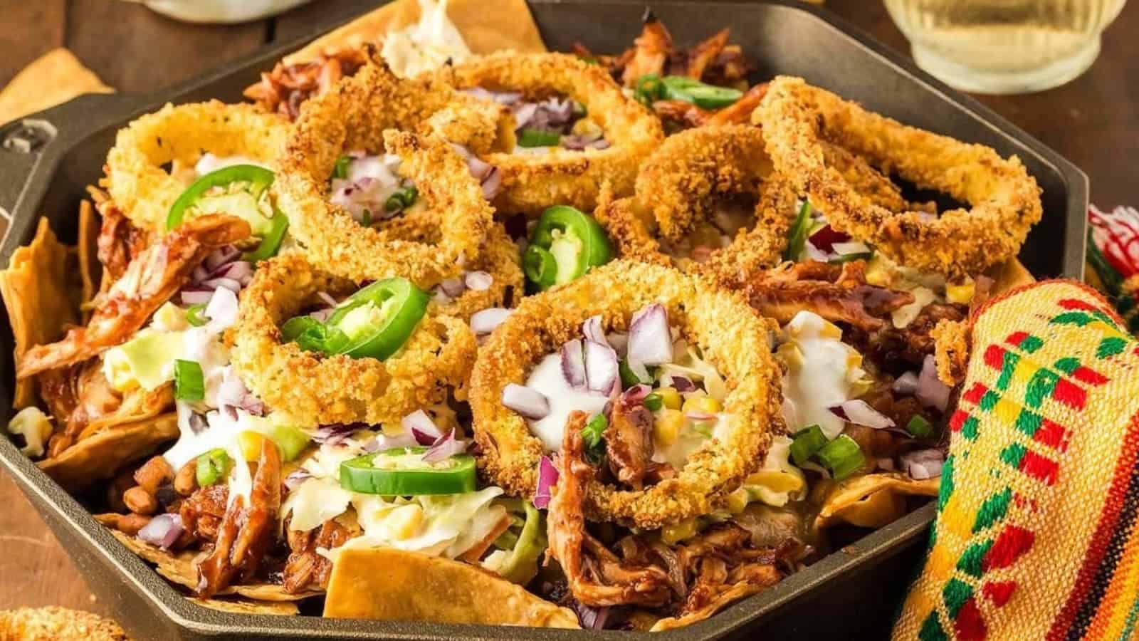 A skillet of nachos topped with pulled pork, onion rings, jalape&ntilde;os, sour cream, and chopped onions.