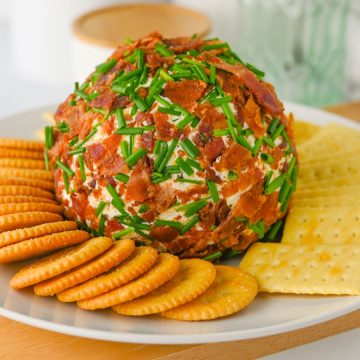 A cheese ball coated in bacon bits and chives, served with round and square crackers on a white plate.