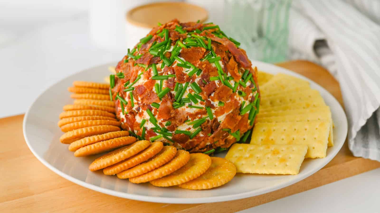 A cheese ball coated in bacon bits and chives, served with round and square crackers on a white plate.