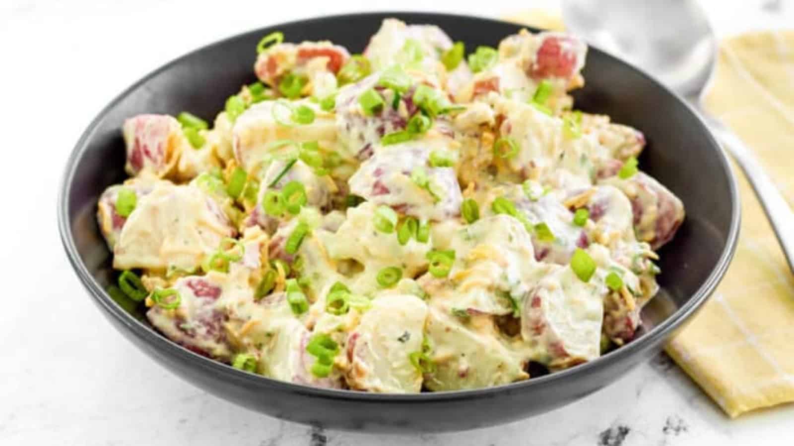 A black bowl of creamy potato salad topped with chopped green onions, with a spoon and napkin beside it.