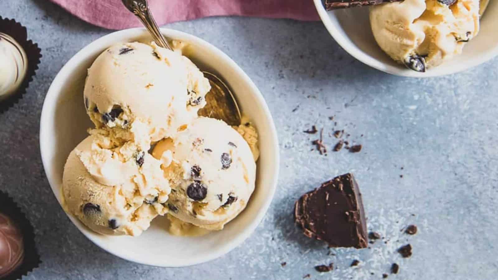 Three scoops of chocolate chip ice cream in a bowl with a spoon, next to chocolate pieces on a gray surface.