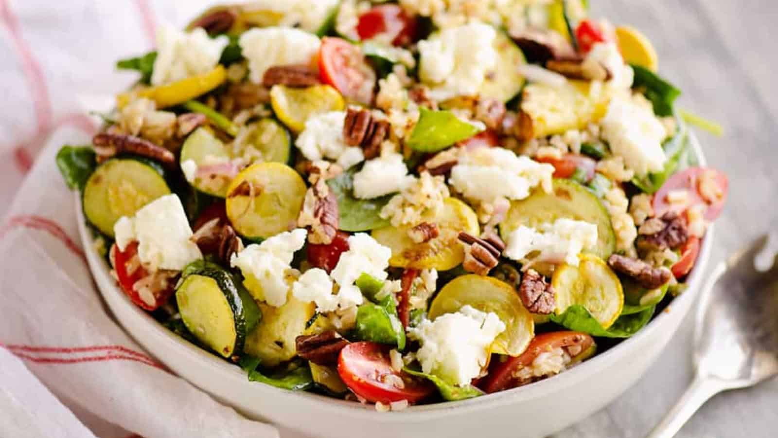 A bowl of salad with zucchini, cherry tomatoes, feta cheese, pecans, and leafy greens.