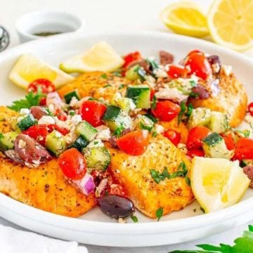 Grilled salmon fillets topped with a fresh cucumber, tomato, olive, and feta salad, garnished with lemon wedges.