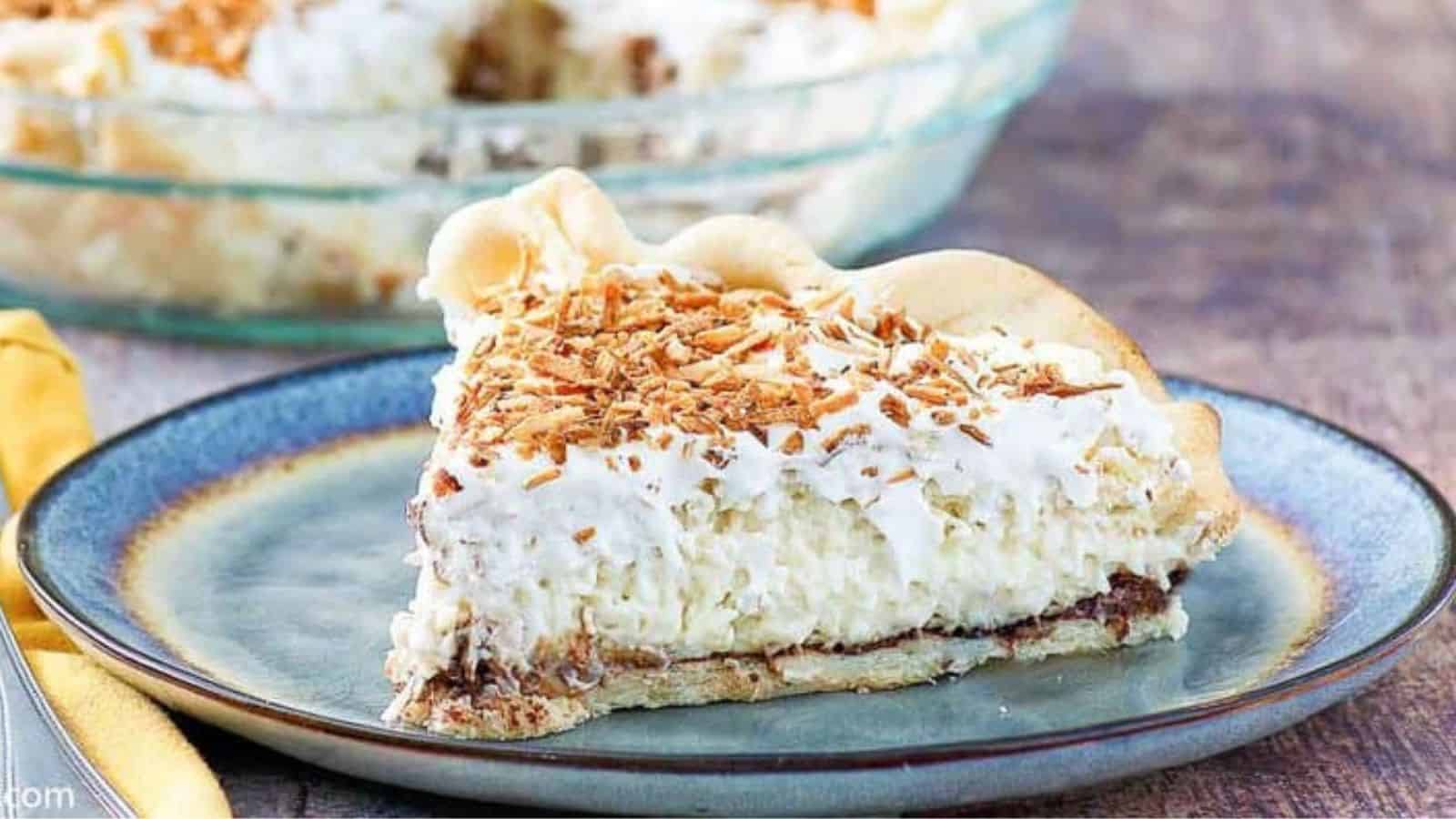 A slice of creamy pie topped with toasted coconut sits on a blue plate, with the whole pie in the background.