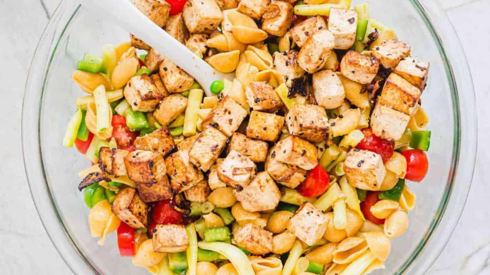 A bowl of pasta salad with grilled tofu, cherry tomatoes, green bell peppers, and a white serving spoon.