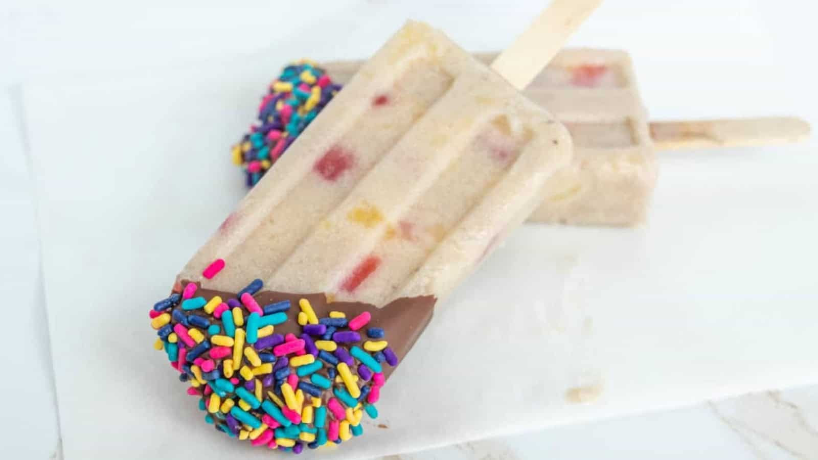 Two creamy popsicles with colorful sprinkles and chocolate on one end, resting on a white surface.