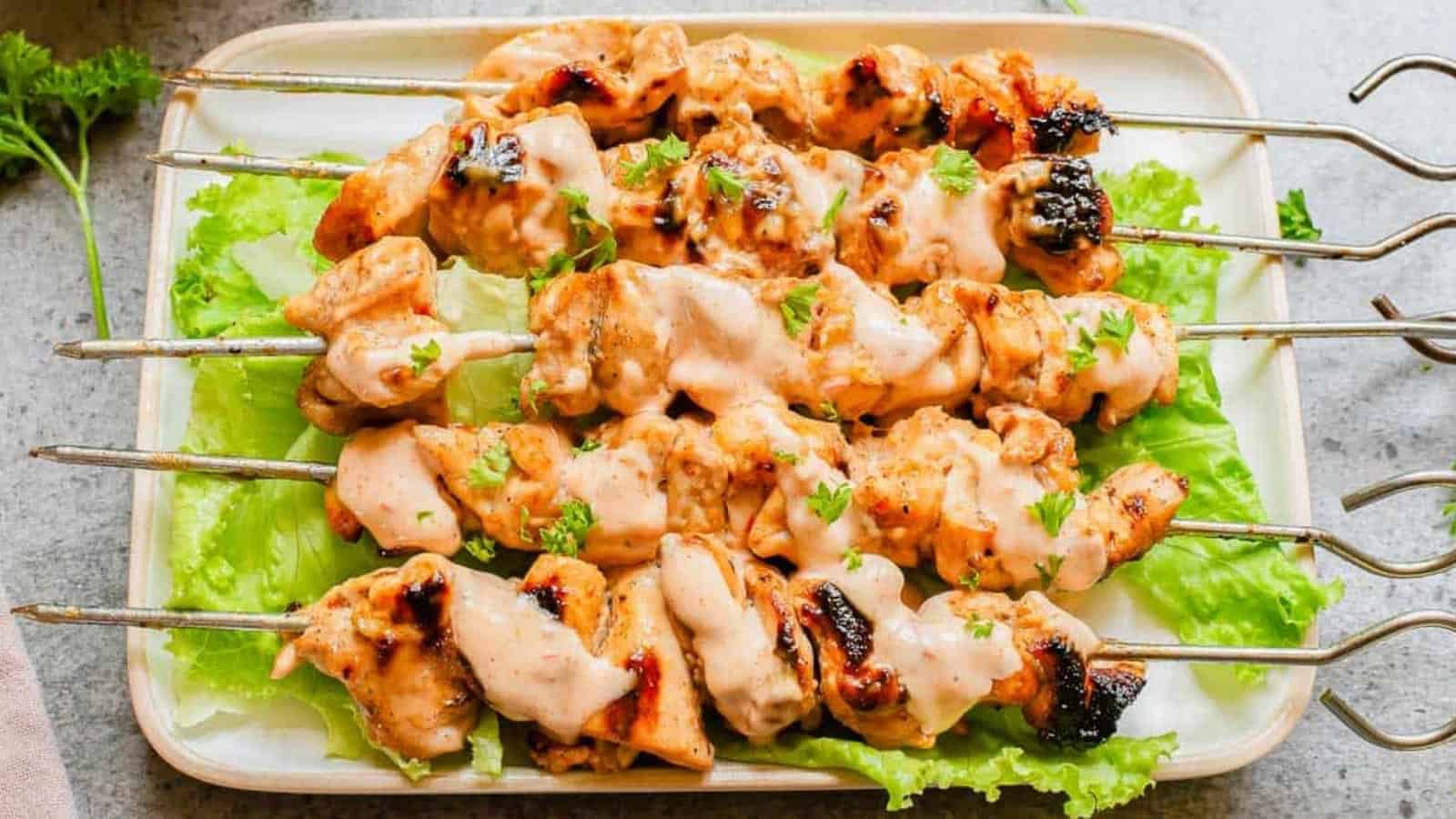 Grilled chicken skewers with creamy sauce, served on lettuce leaves on a rectangular white plate.