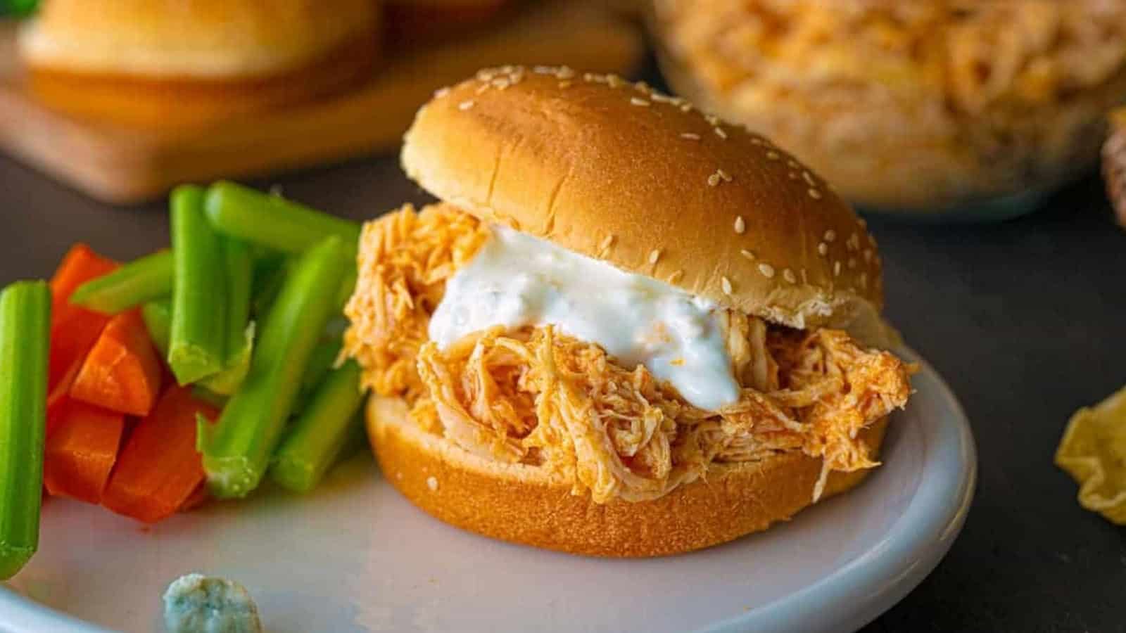 A pulled chicken sandwich with ranch sauce, served with celery and carrot sticks on a white plate.
