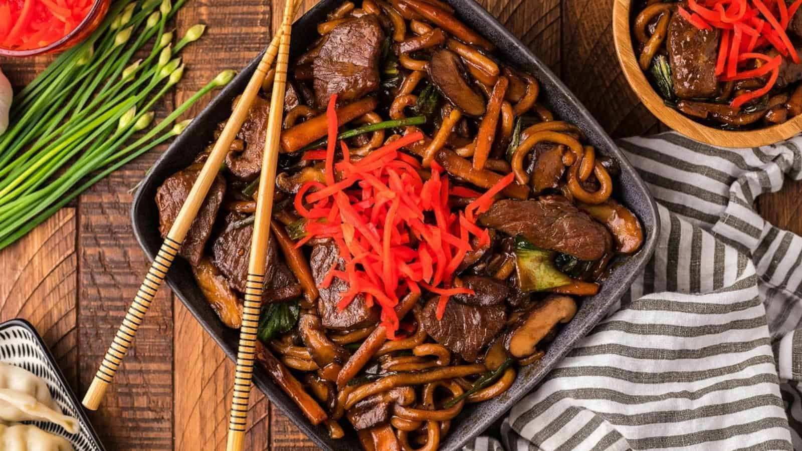 Square bowl of beef stir-fried noodles with red pickled ginger, chopsticks, and vegetables on a wooden table.