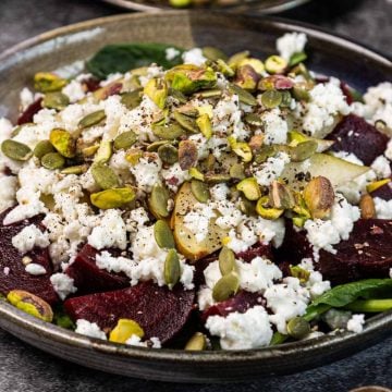 Salad with beetroot, crumbled feta, pumpkin seeds, pistachios, spinach, and pears on a dark plate.
