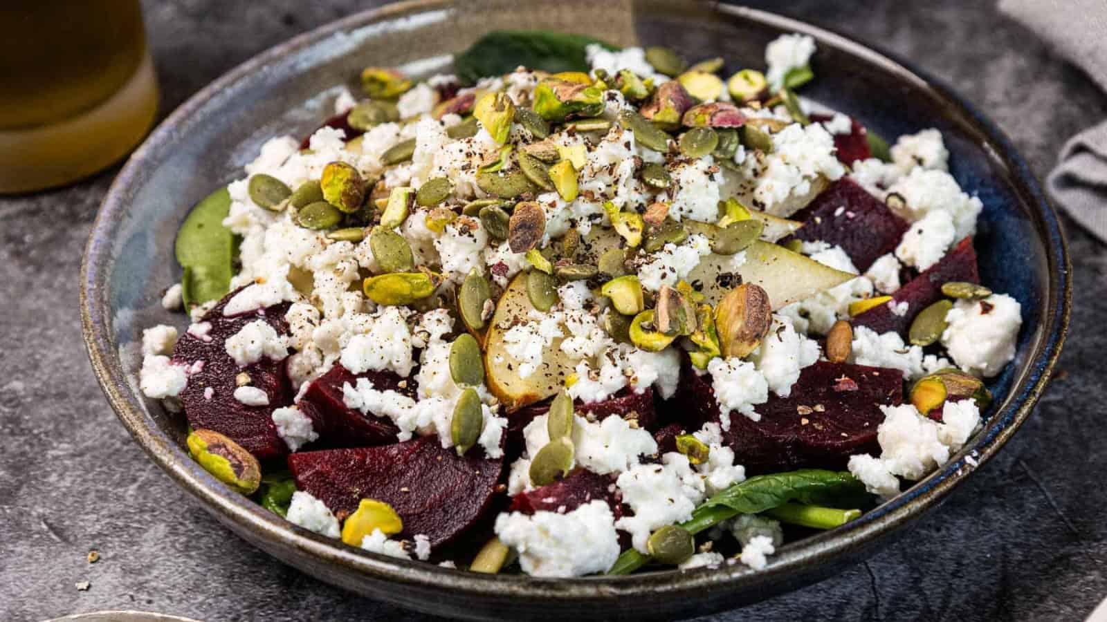 Salad with spinach, beetroot, crumbled feta, pear slices, and mixed seeds served in a dark bowl.