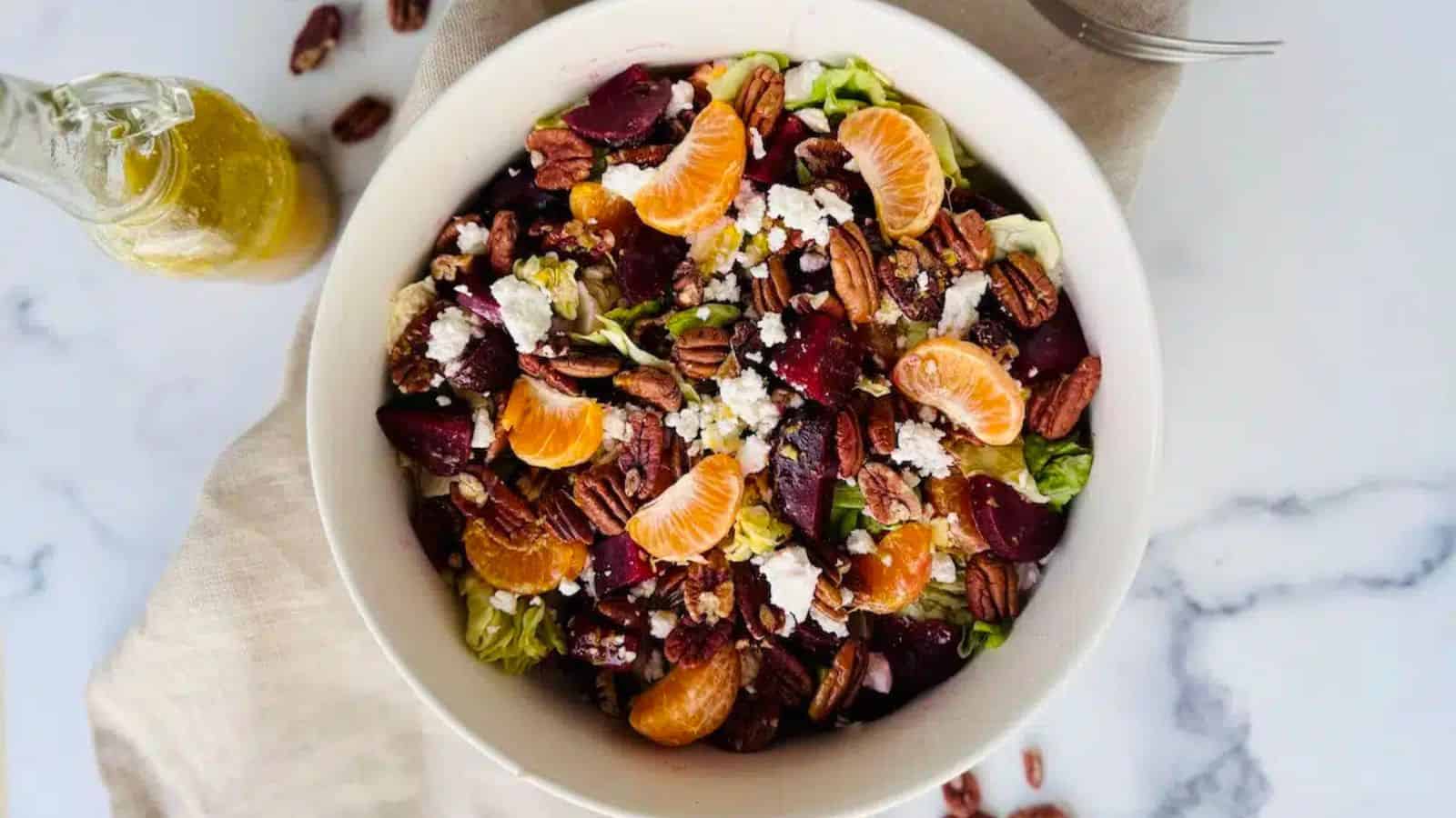 Bowl of salad with mandarin slices, pecans, feta cheese, beets, and greens on a white surface.