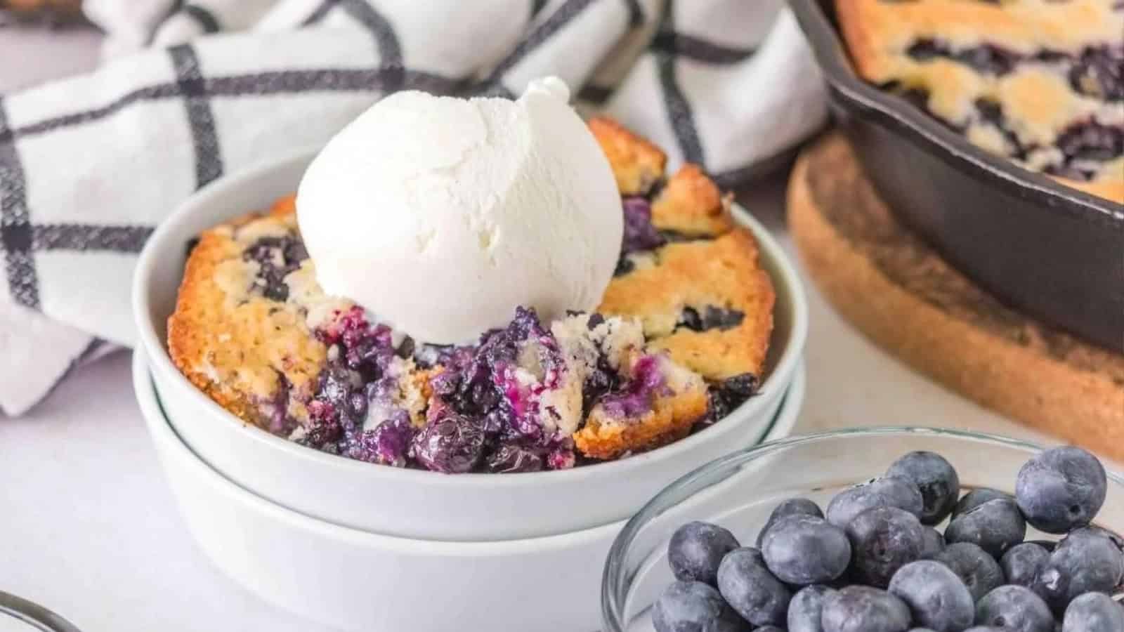 A bowl of blueberry cobbler topped with a scoop of vanilla ice cream, with fresh blueberries beside it.