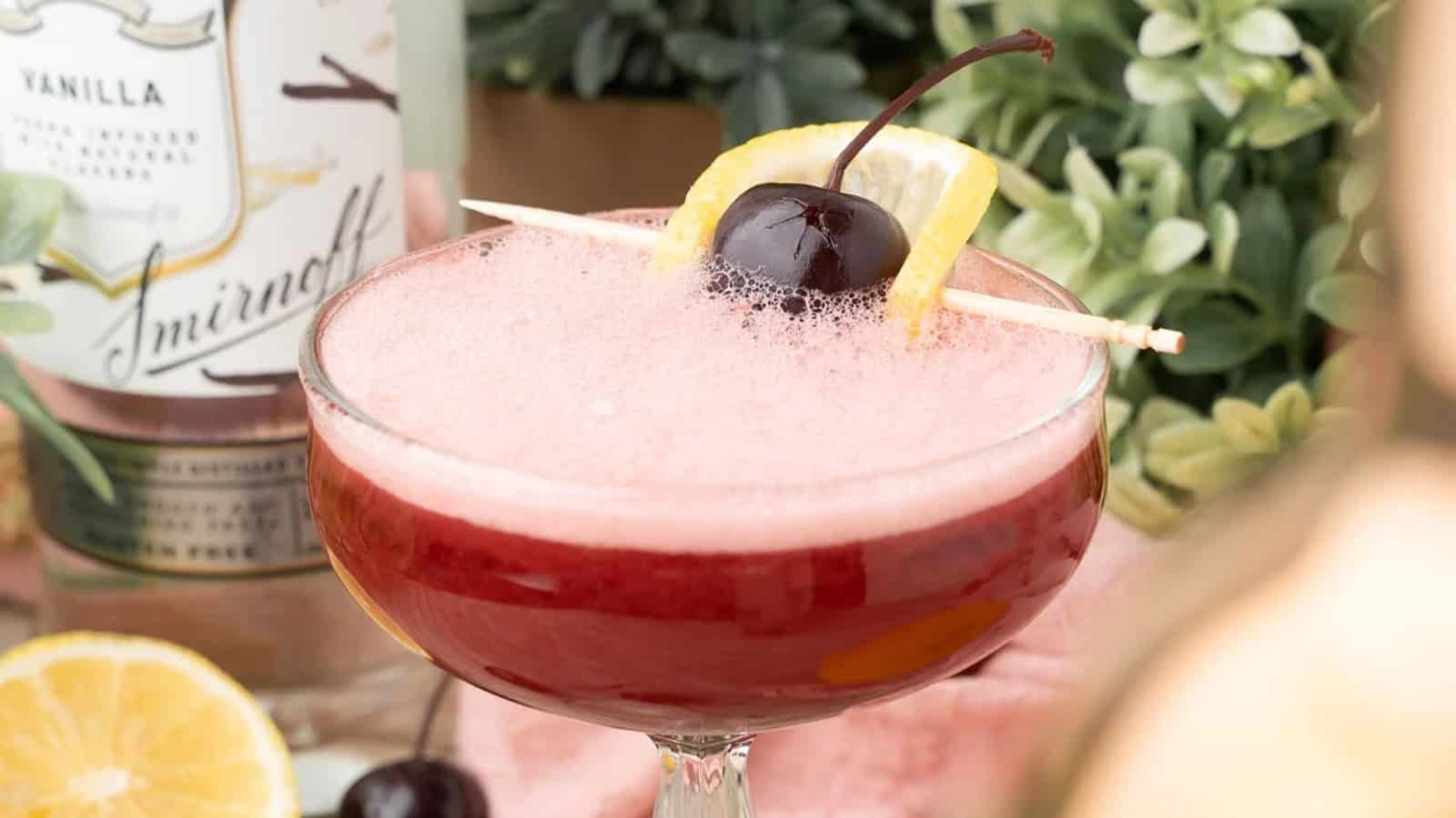 A pink cocktail garnished with a cherry and lemon slice on a toothpick, with a bottle and greenery in the background.