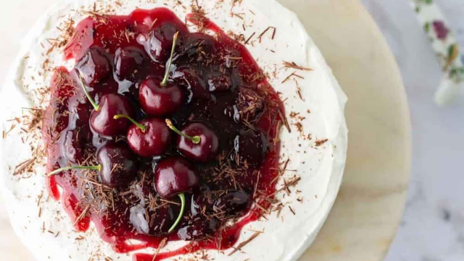 A cake topped with whipped cream, cherry sauce, whole cherries, and chocolate shavings on a light surface.