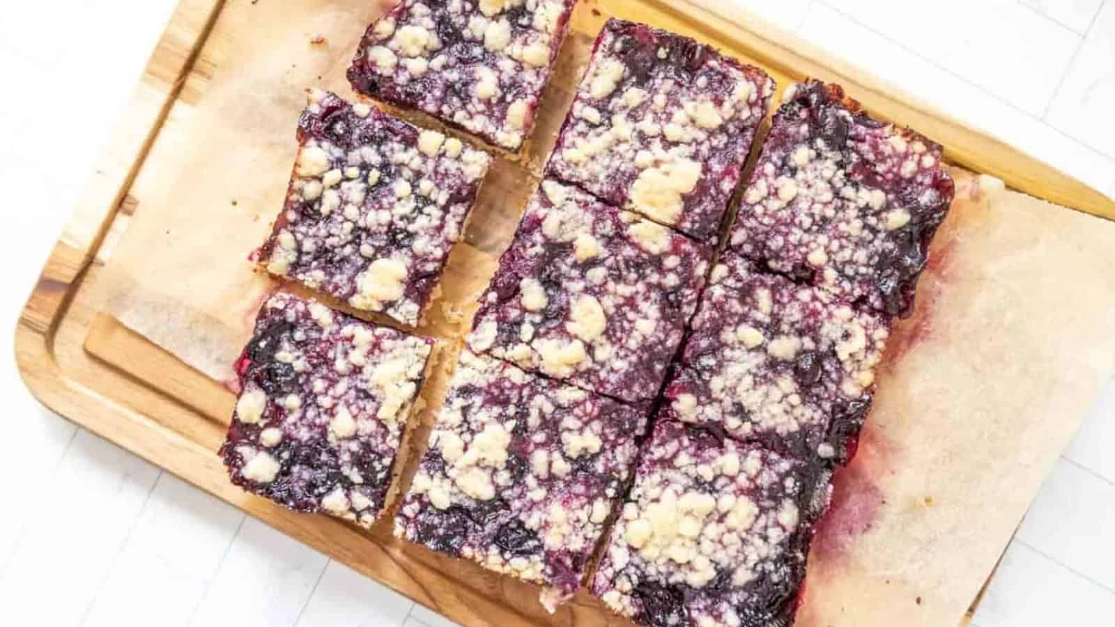 Nine square berry crumble bars on parchment paper atop a wooden cutting board.
