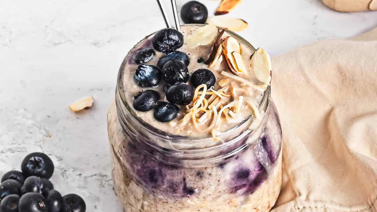 A jar of overnight oats topped with fresh blueberries, sliced almonds, and shredded coconut on a light surface.