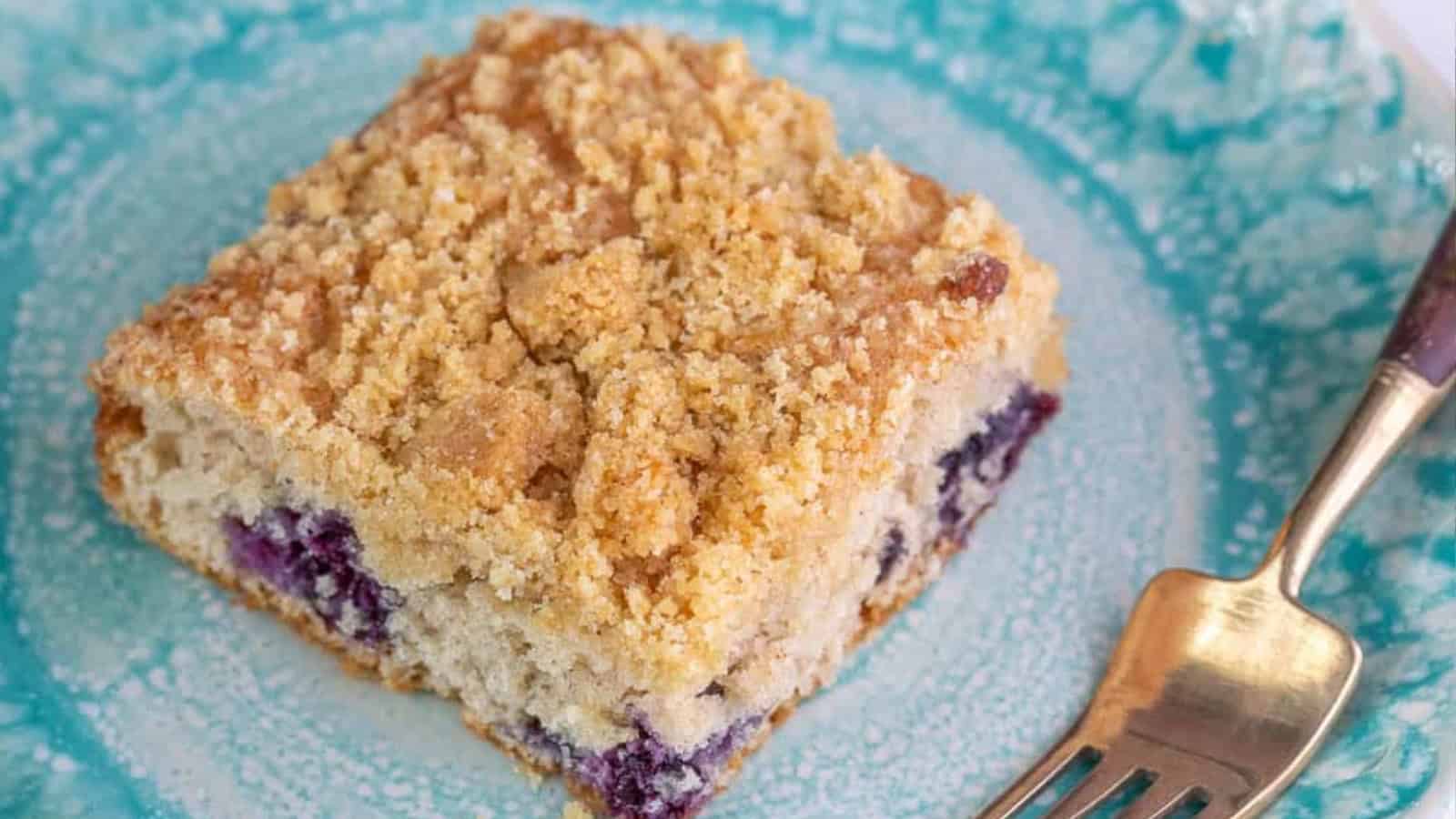 A slice of crumb-topped blueberry coffee cake on a blue plate with a fork.