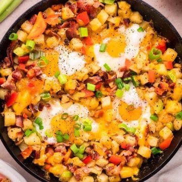 Skillet breakfast hash with eggs, potatoes, diced veggies, bacon, and green onions, garnished with black pepper.