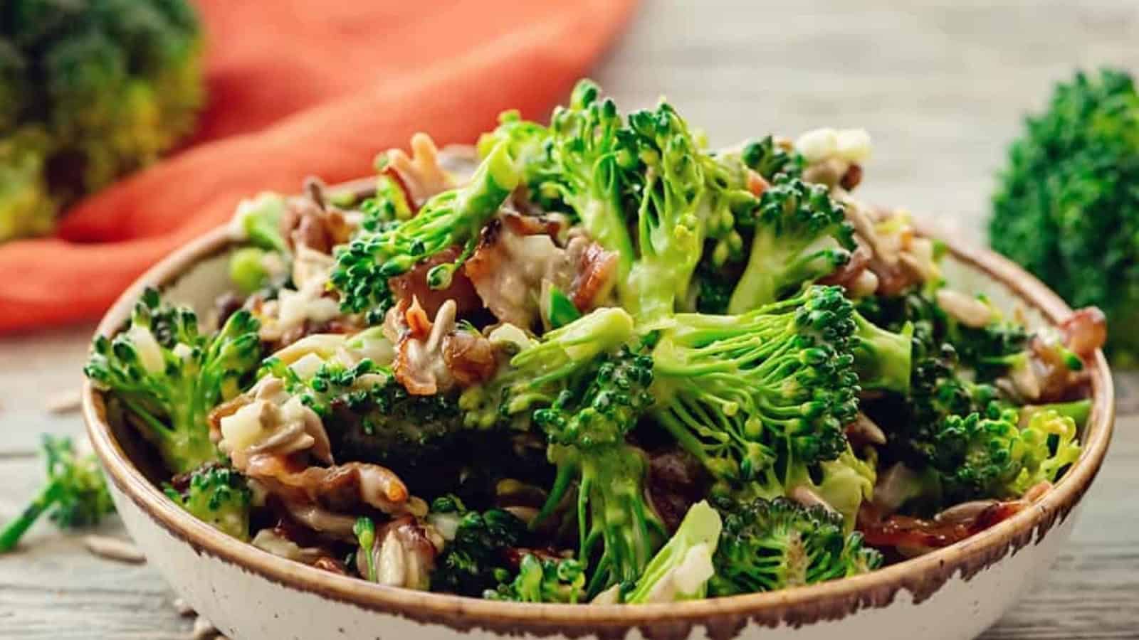 A bowl of fresh broccoli salad with sliced onions, nuts, and a creamy dressing on a light wooden table.
