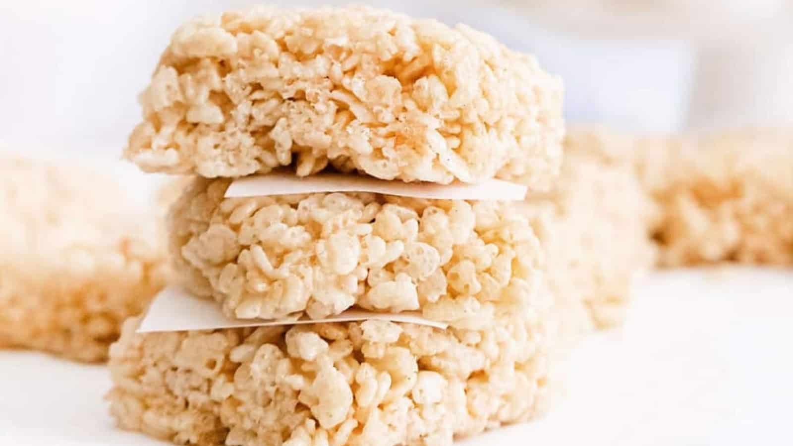 Three rice krispie treats stacked with parchment paper between them, with more treats blurred in the background.