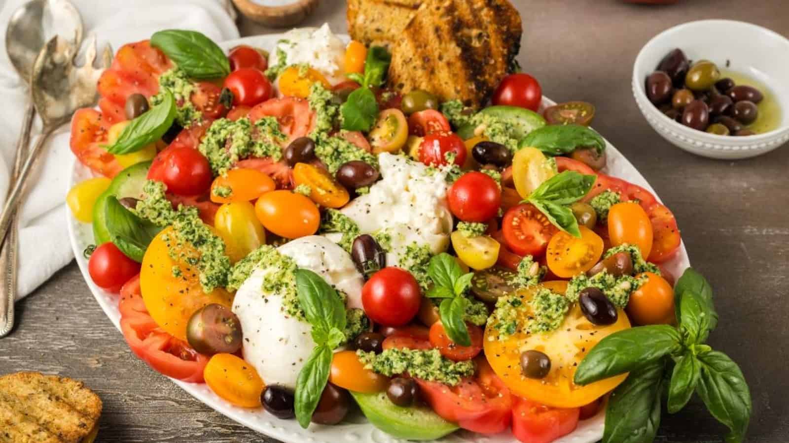 Colorful tomato salad with mozzarella, olives, basil, pesto, and toasted bread on a white platter.