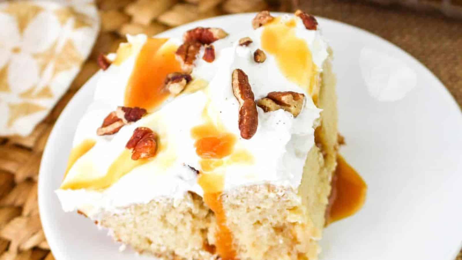A slice of cake on a white plate, topped with whipped cream, caramel sauce, and nuts. The rest of the cake is visible in the background on a woven mat.