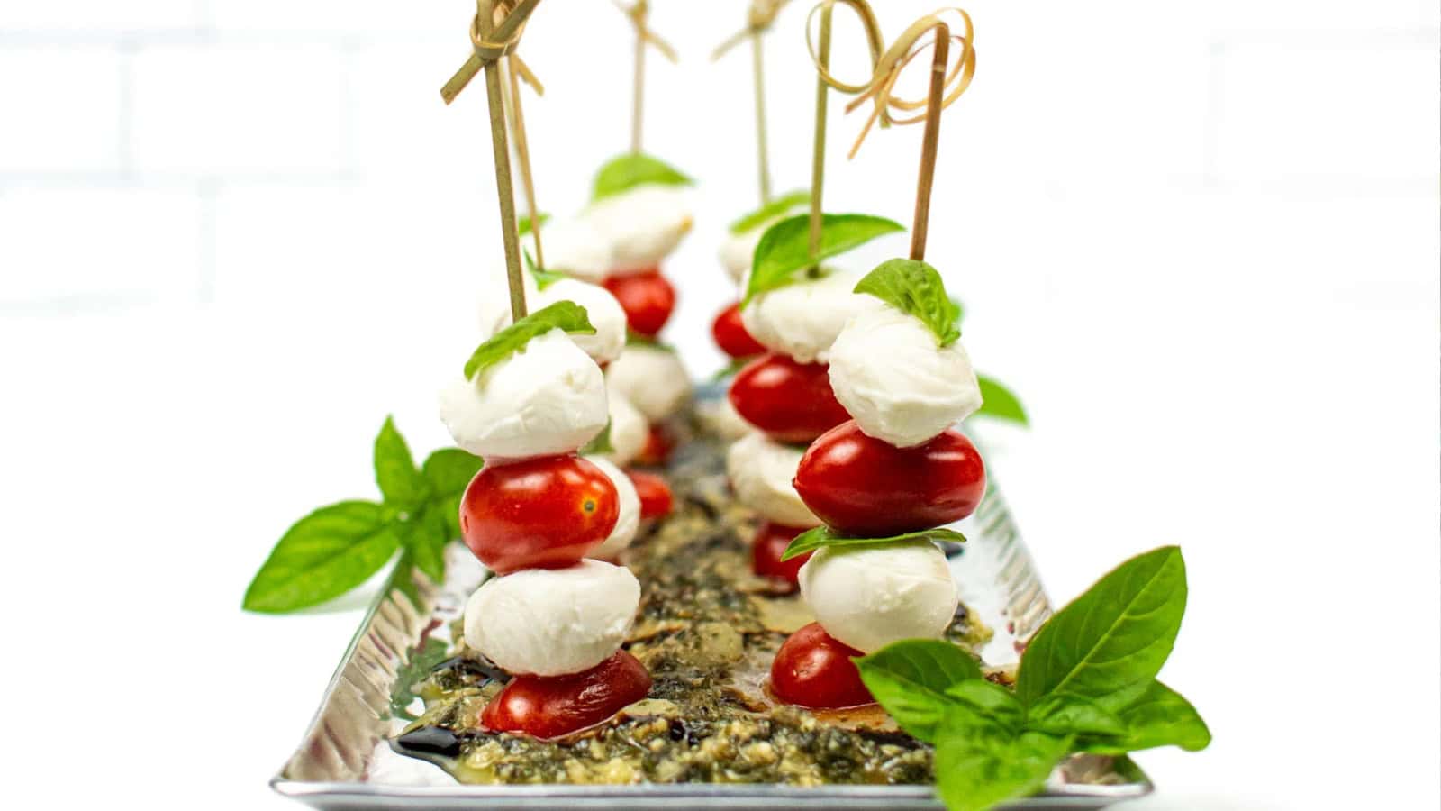 Skewers with mozzarella, cherry tomatoes, and basil, arranged on a platter with pesto and basil leaves.