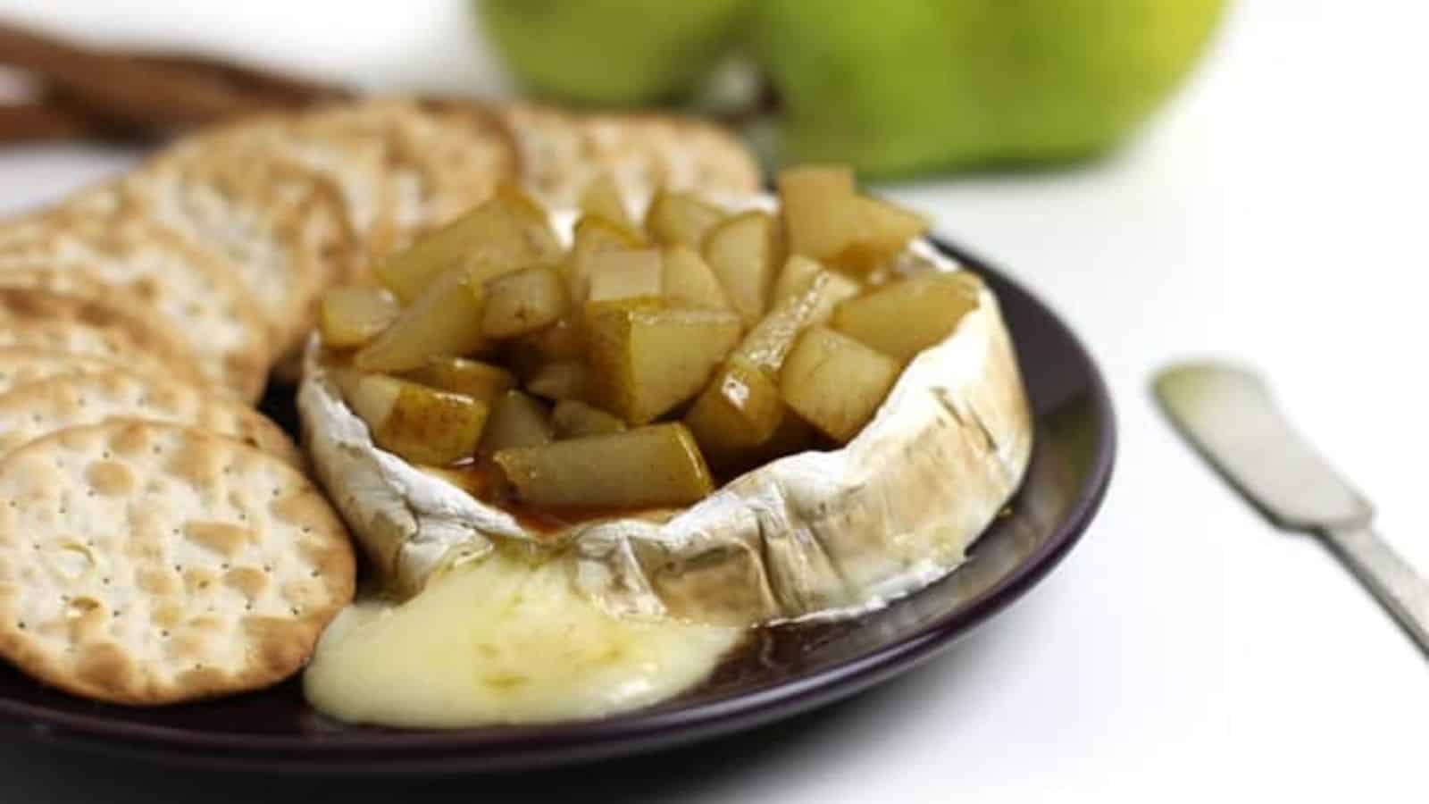 Baked brie topped with diced pears and served with crackers on a dark plate, with cheese melting out.
