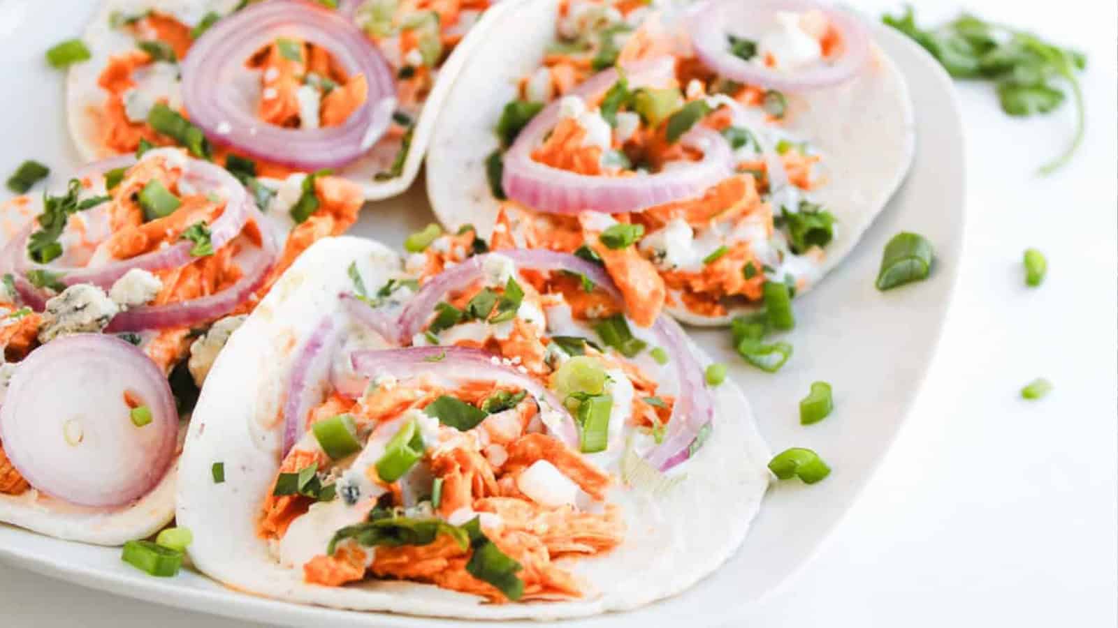 Four tacos filled with shredded meat, sliced red onions, green onions, and creamy sauce on a white plate.