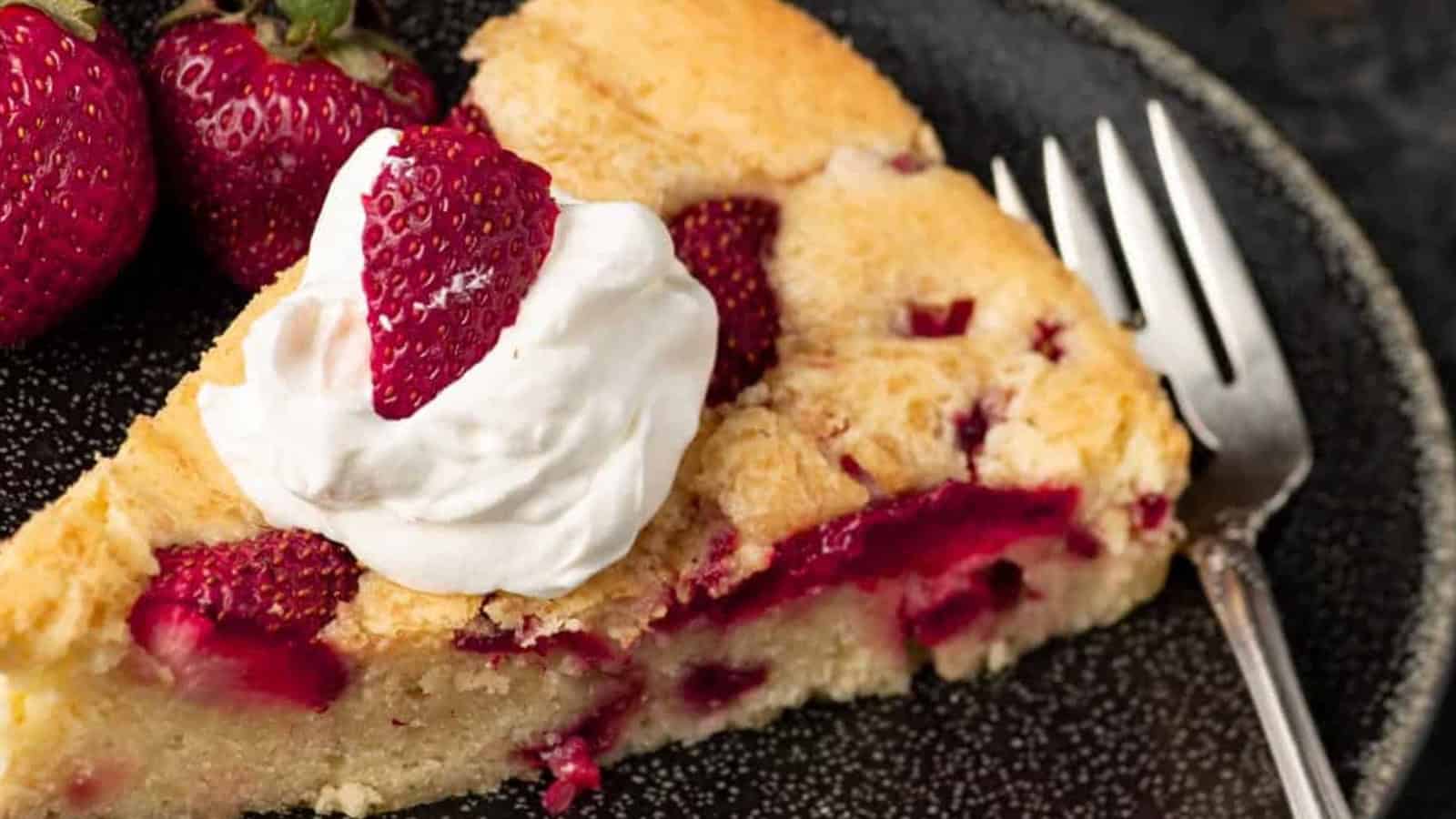A slice of strawberry cake with whipped cream and strawberries, served on a black plate with a fork.