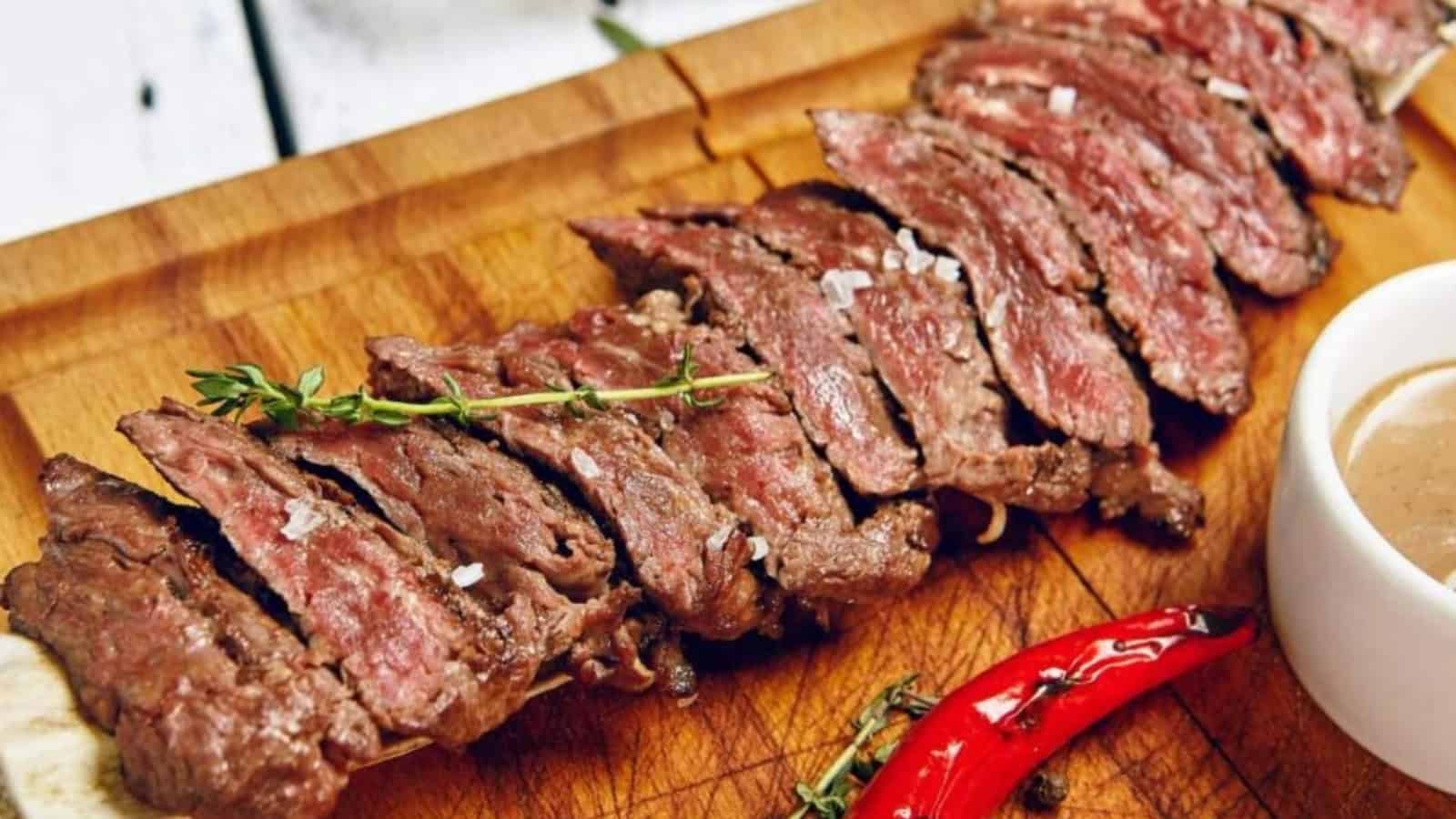 Sliced medium-rare grilled steak garnished with thyme and a red chili pepper on a wooden cutting board.