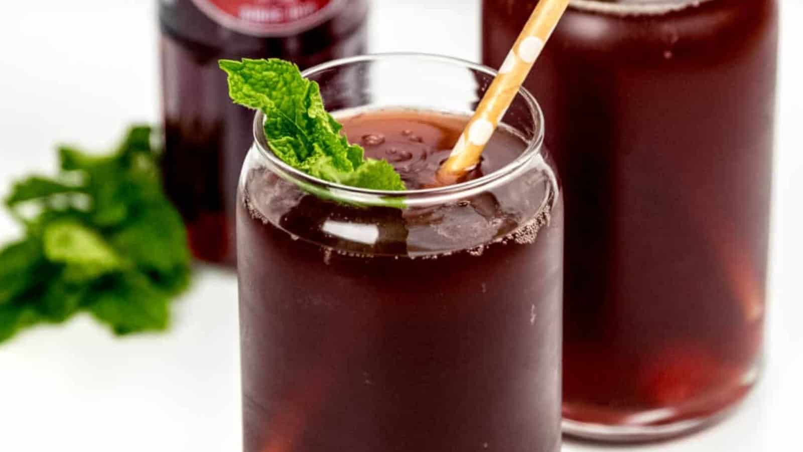 A glass of dark red drink with ice, mint garnish, and a straw, with bottles and mint leaves in the background.
