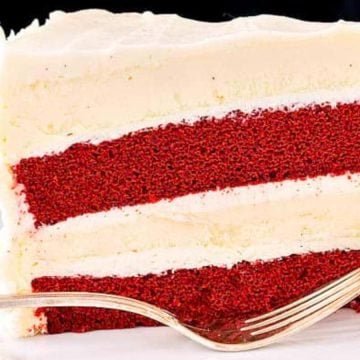 A slice of red velvet cake with cream cheese frosting, served with a swirl of whipped cream and a fork.