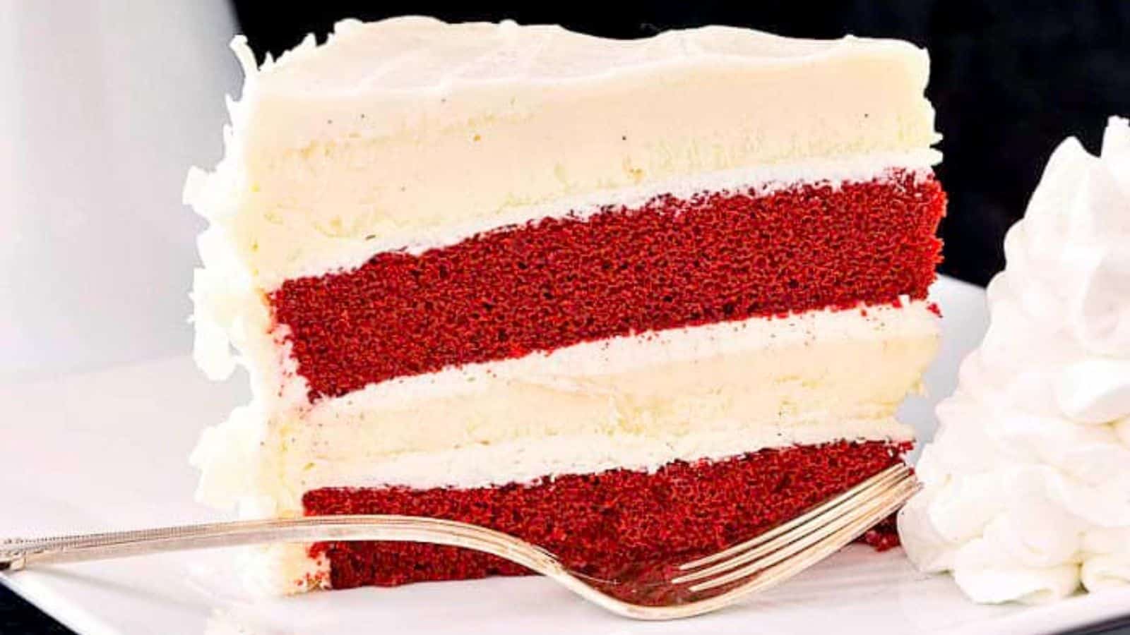 A slice of red velvet cake with cream cheese frosting, served with a swirl of whipped cream and a fork.