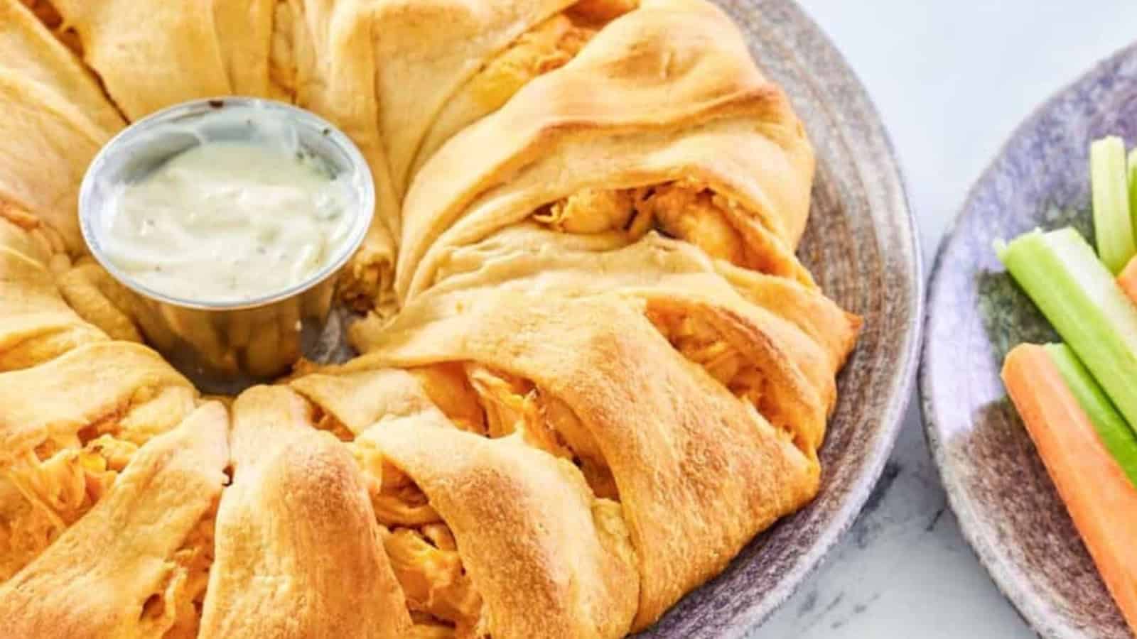 A golden crescent roll ring filled with chicken, served with ranch dip and a side of celery and carrot sticks.