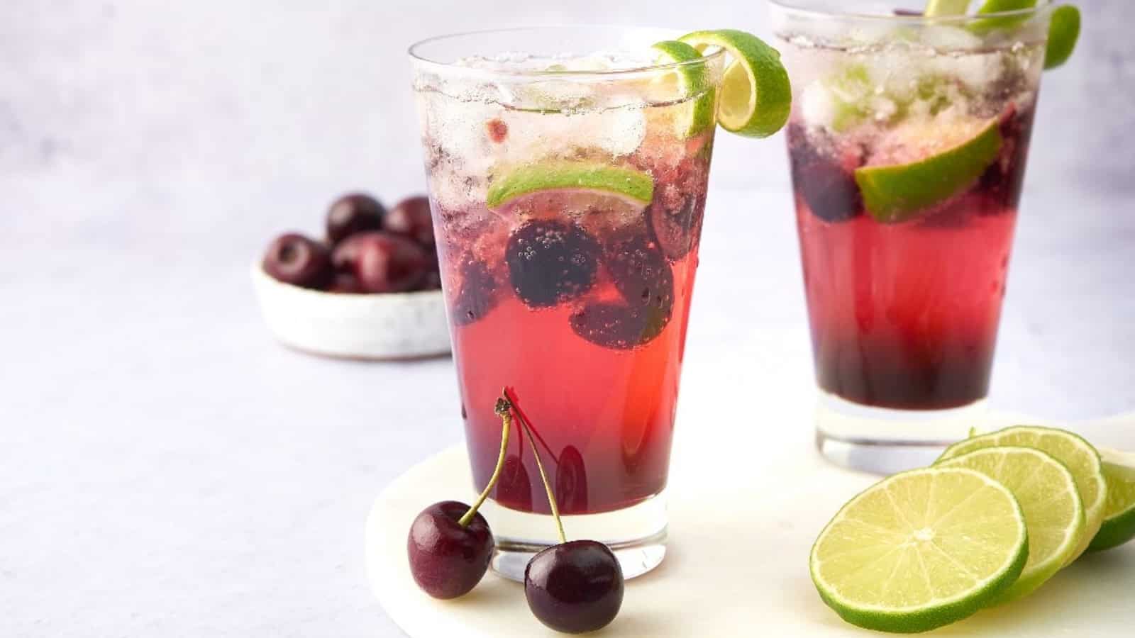 Two glasses of cherry lime drink with ice, lime slices, and fresh cherries, with extra fruit on the side.
