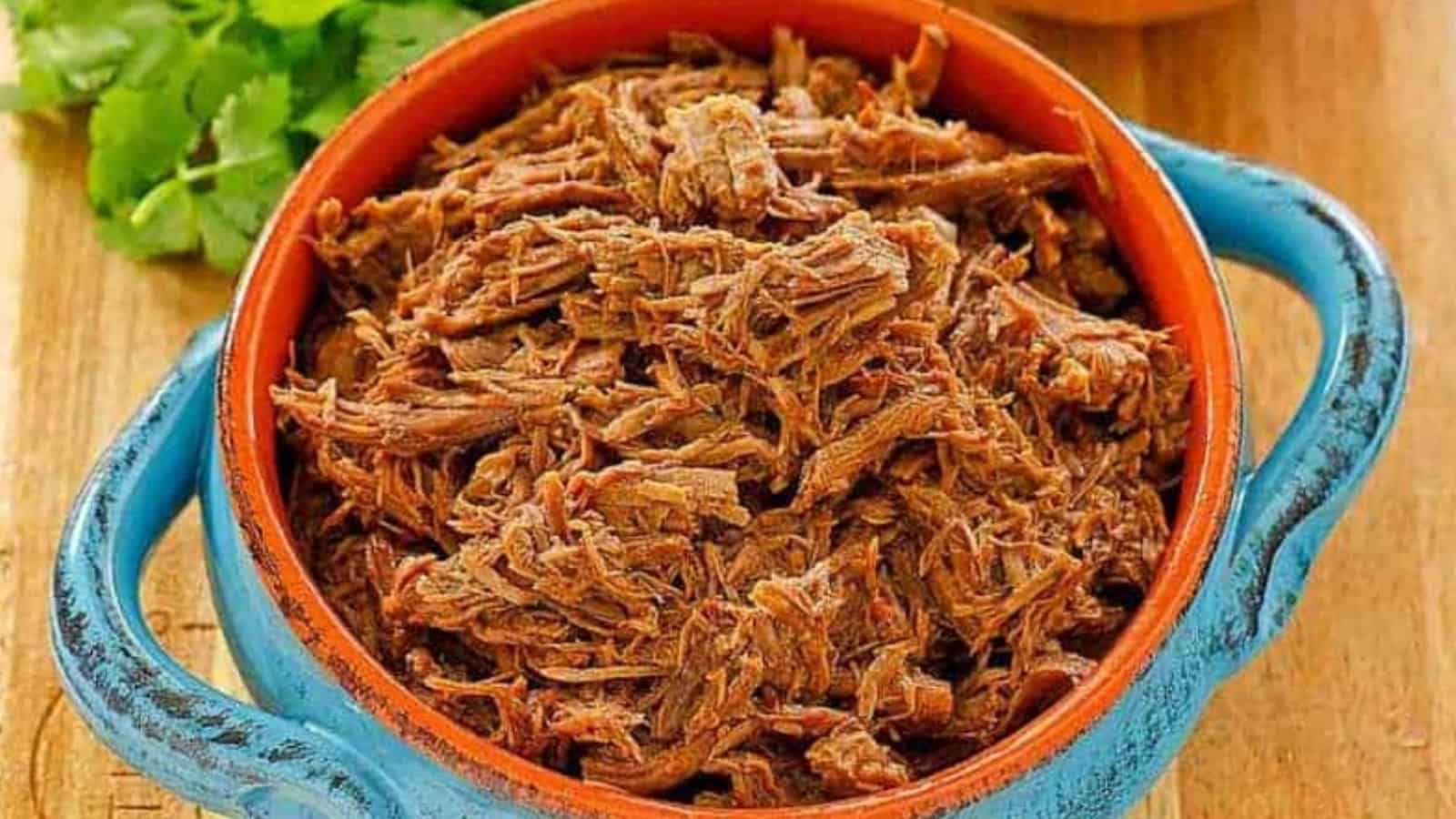 Shredded beef barbacoa served in a rustic blue and brown bowl on a wooden table with cilantro nearby.