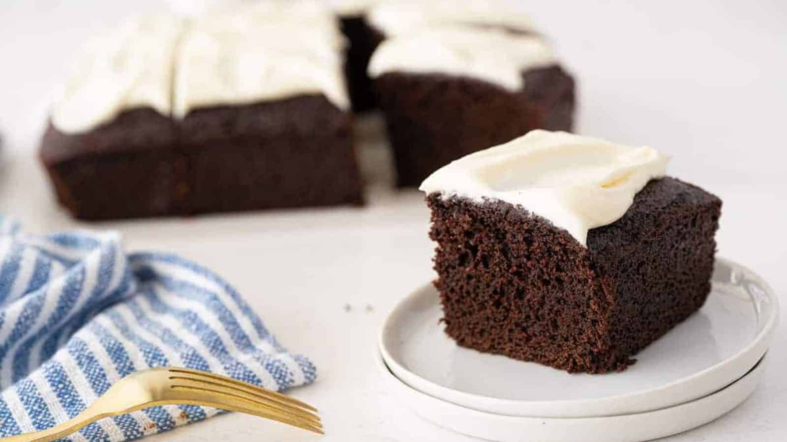 A slice of chocolate cake with white frosting on a plate, with more cake in the background.