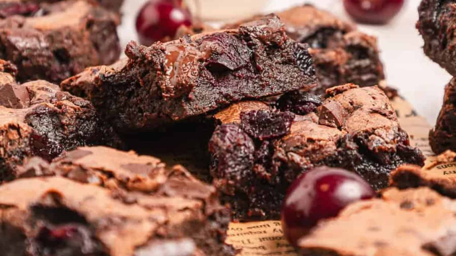 Chocolate brownies with chunks of cherry and whole cherries scattered around on a brown surface.