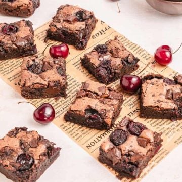 Brownie squares with cherries scattered on vintage paper, surrounded by cups of coffee and a plate.