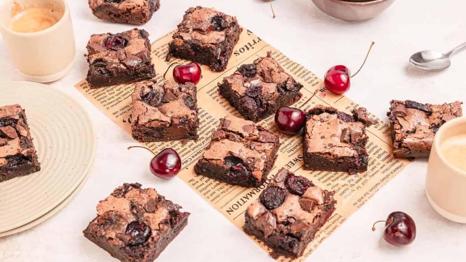 Brownie squares with cherries scattered on vintage paper, surrounded by cups of coffee and a plate.