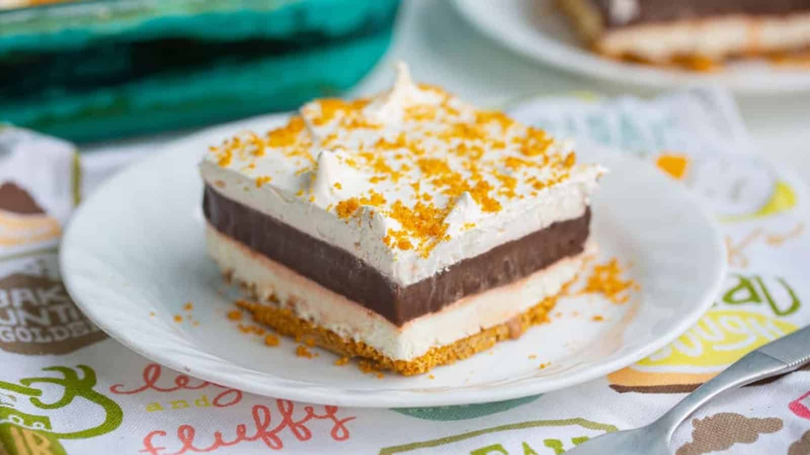 A slice of layered dessert with whipped topping, chocolate, and a crumbly crust on a white plate.