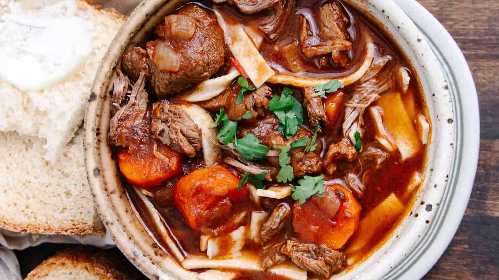 A bowl of beef stew with carrots, noodles, and parsley, served with slices of bread.