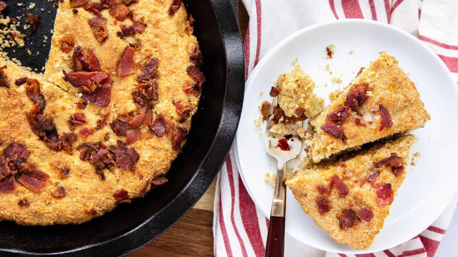 A skillet of cornbread topped with bacon beside a plate with two sliced pieces and a fork.