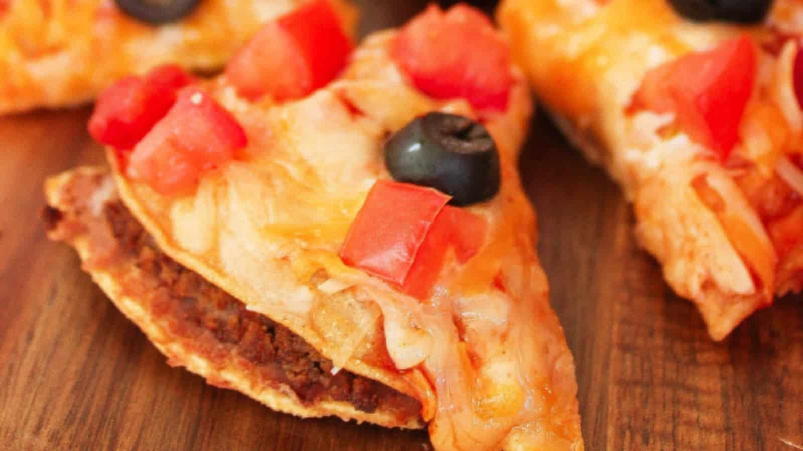 A slice of taco pizza topped with cheese, tomatoes, onions, and a black olive on a wooden surface.