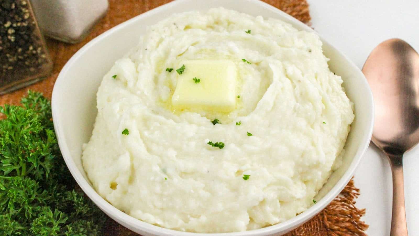 Bowl of mashed potatoes with a pat of butter on top, garnished with parsley. A spoon and a sprig of parsley are next to the bowl.