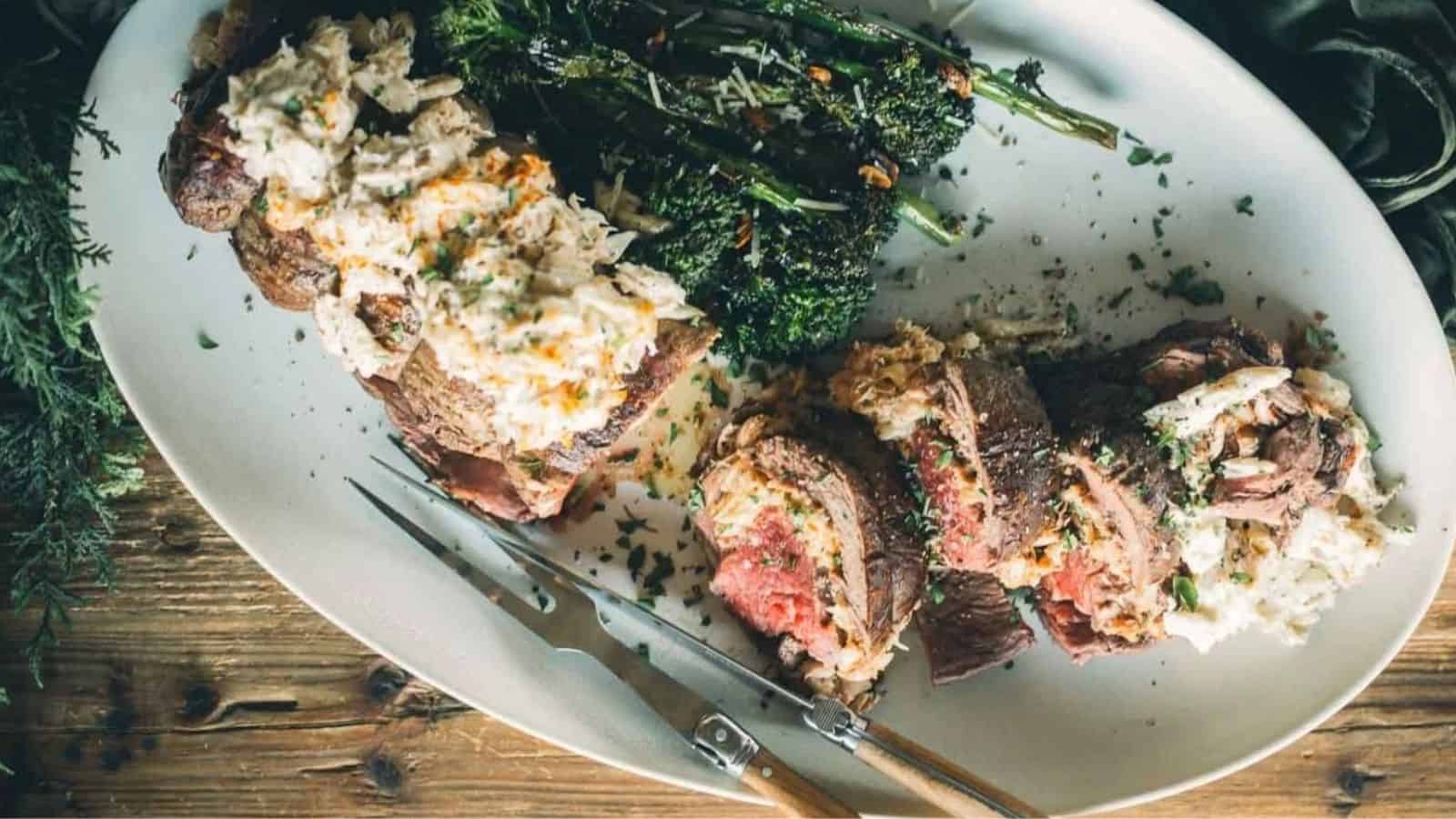 Sliced steak topped with creamy sauce, served with broccolini on a white plate with carving utensils.