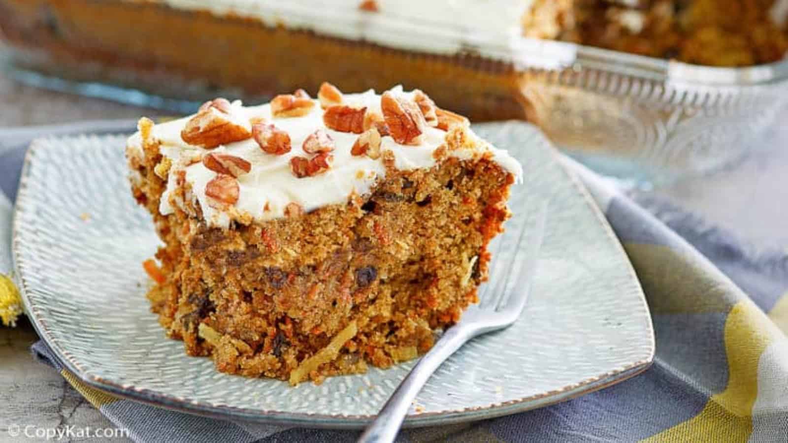 A slice of carrot cake with cream cheese frosting and chopped pecans on a plate with a fork.