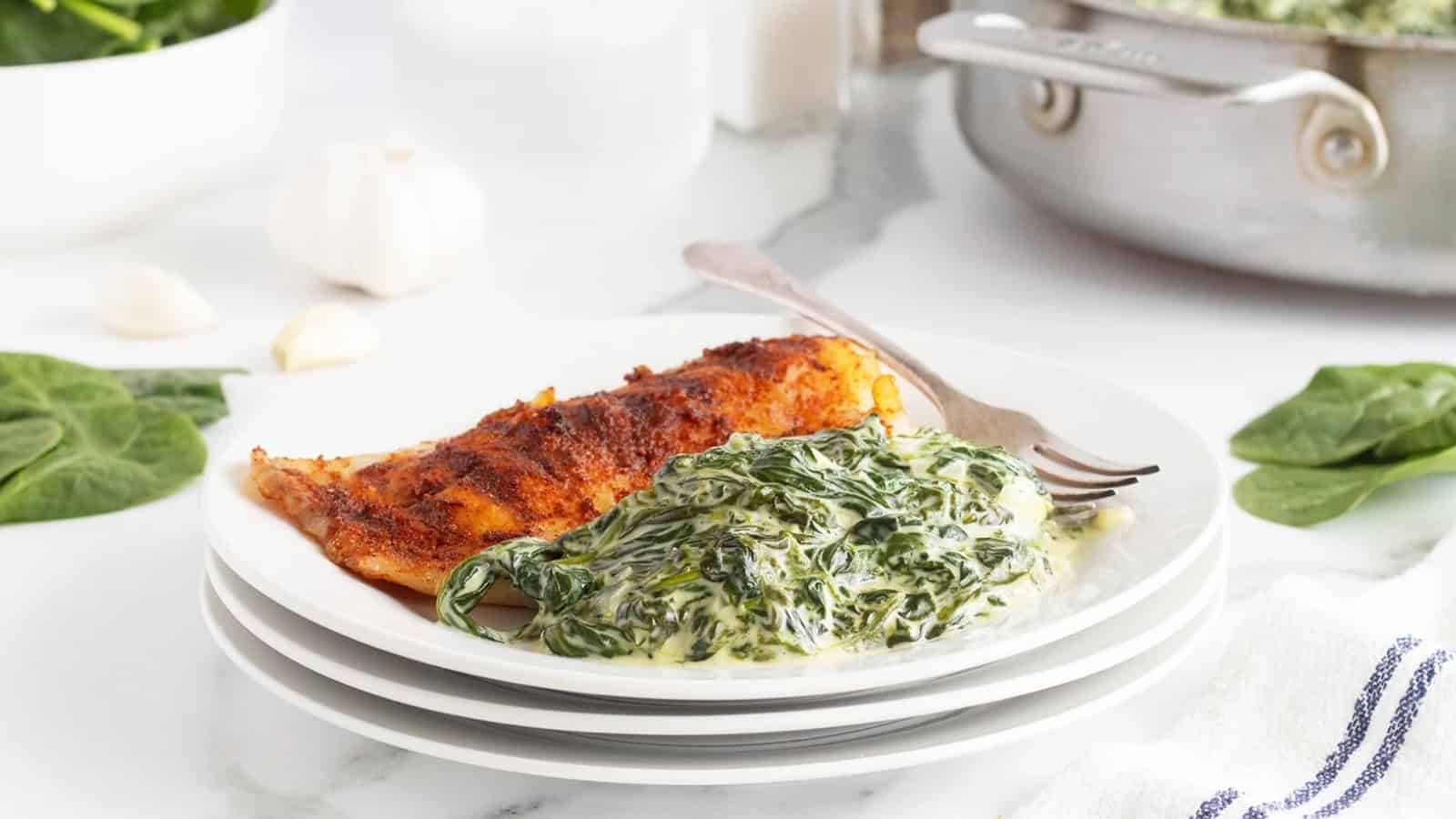 A white plate with creamed spinach and a piece of seasoned fish, with a fork on the side.